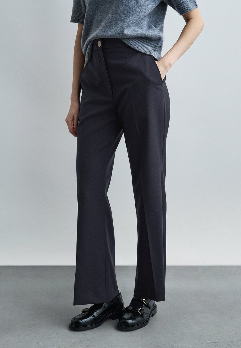 Une personne debout, une main dans la poche, portant un pantalon large marine, un pull gris à manches courtes, des mocassins brogues noirs, sur un sol et un fond gris.