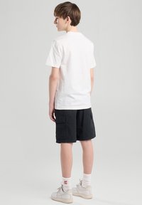 Valkoinen puuvillapaita, takanäkemys, yhdistetty mustiin cargo-shorteihin. Mallilla on valkoiset lenkkarit punaisilla logosukilla. Yksinkertainen tausta.