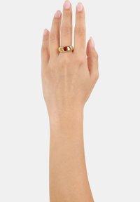 Mano con uñas pulidas que lleva un anillo de oro con una gema en forma de corazón rojo en el dedo medio, sobre un fondo claro y liso.