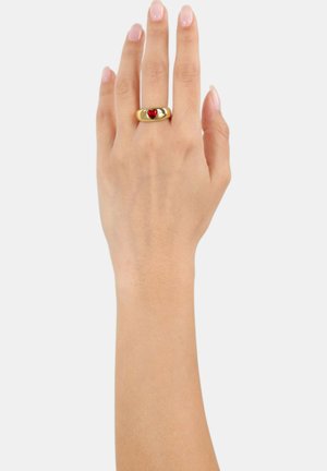 Mano con uñas pulidas que lleva un anillo de oro con una gema en forma de corazón rojo en el dedo medio, sobre un fondo claro y liso.