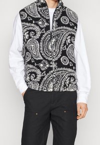 Gilet in pile con motivo paisley bianco e nero, colletto alto e zip frontale, abbinato a una camicia bianca e pantaloni neri.