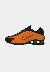 Sapatilha desportiva Nike com um upper preto e detalhes em laranja vibrante em material sintético. Apresenta amortecimento Shox e um design perfurado para respirabilidade.