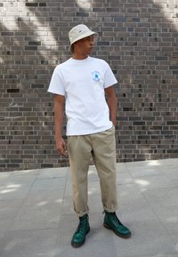 White cotton t-shirt with blue graphic, beige trousers, and green leather boots. The outfit features a bucket hat, completing the casual look.