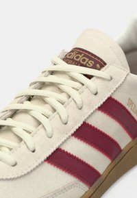 Beige ruskindssneakers med røde gummistriber og lyse snørebånd. Har en mørkerød label på tungen med teksten "Adidas Handball".