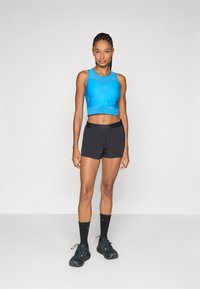 Plavi atletski crop top glatke teksture, uparen s crnim šorcem. Model nosi crne čarape i tenisice, stojeći na neutralnoj pozadini.