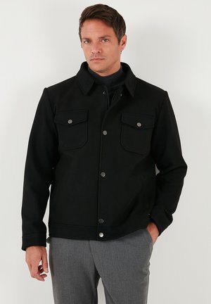 Vastag anyagból készült fekete dzseki, galléros nyakkivágással, elől gombos záródással, valamint dupla mellzsebekkel, melyek fém patentokkal vannak ellátva.