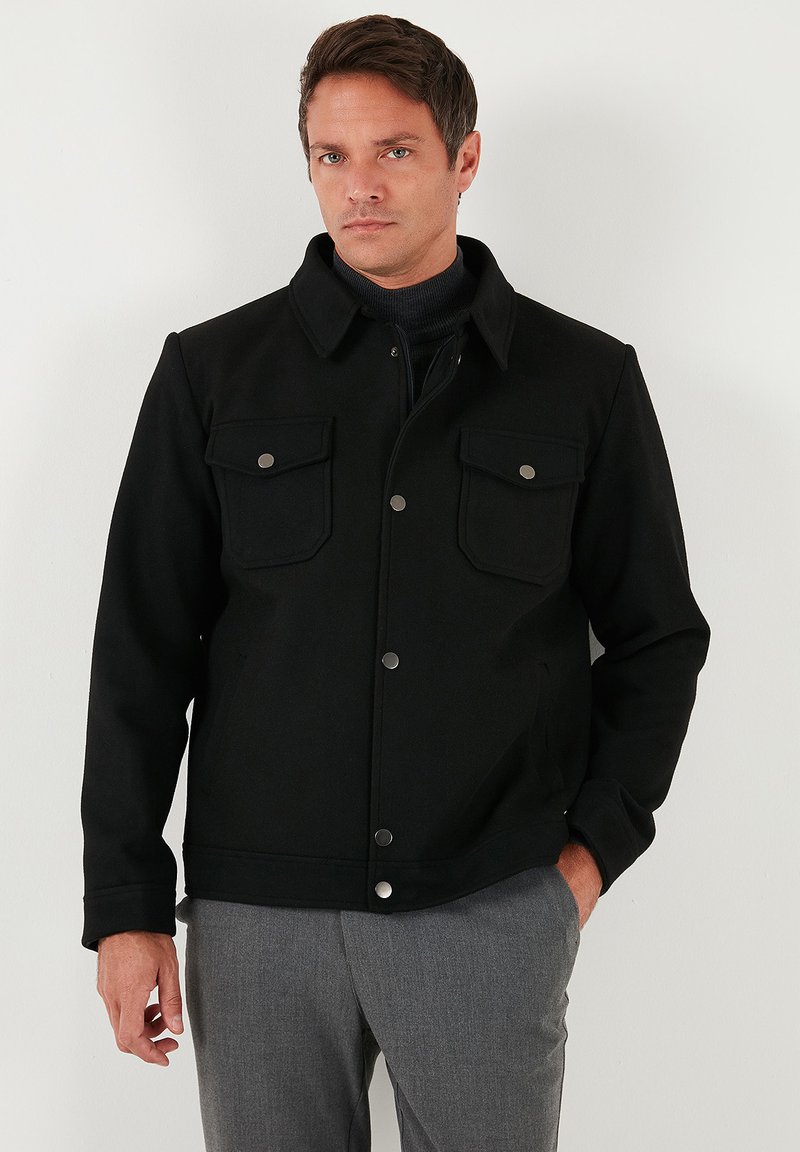 Juoda striukė iš storesnės medžiagos, su apykakle, priekyje užsegama sagomis ir dviguba krūtinės kišenė su metaliniais užsegimais.