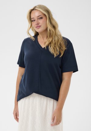 Blondynka z luźno ułożonymi falami włosów nosi granatowy top z krótkim rękawem i dekoltem w serek oraz białą teksturowaną spódnicę na jednolitym tle.