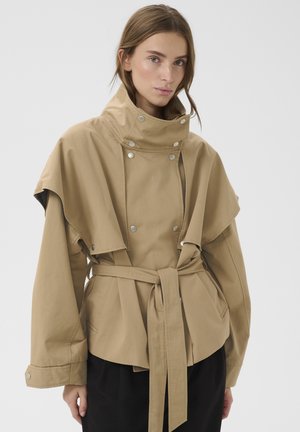 Femeie purtând o jachetă bej cu guler înalt și curea, cu nasturi argintii, și pantaloni negri, stând în fața unui fundal alb simplu.