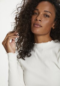 Haut à manches longues blanc côtelé avec un col ondulé et un tissu texturé. Le modèle a des cheveux bouclés et touche ses cheveux d'une main.