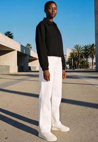 Vrouw staat buiten en draagt een zwarte jas, witte wijde broekspijpen en witte sneakers, met palmbomen en betonnen constructies op de achtergrond.