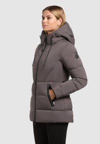 Nais, kes kannab halli pehmendatud talvejakk, millel on kõrge krae, kapuuts, mustad lukuga taskud ja mustad püksid, seistes ühtlase tausta ees.