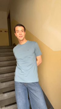 Jeune homme en t-shirt bleu clair et jeans, debout sur un escalier aux murs beiges, souriant, les mains derrière le dos.