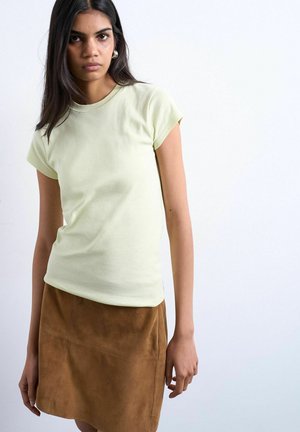 Woman with long dark hair wearing light green fitted t-shirt and brown suede skirt against a plain white background.