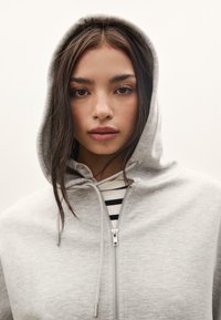 Světle šedá mikina na zip s kapucí na šňůrku, která má hladkou texturu a stříbrný zip. Pod ní je vidět pruhovaná košile.