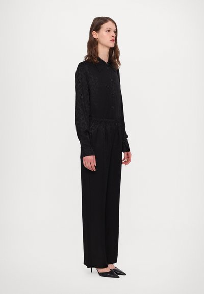 Zwarte jumpsuit met lange mouwen van textuurstof, knoopsluiting aan de voorkant en een elastische tailleband. Gecombineerd met spitse zwarte hakken.