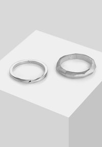 Due anelli d'argento: uno liscio e sottile, l'altro texturizzato con bordi angolari. Entrambi sono rotondi, in mostra un design minimalista.