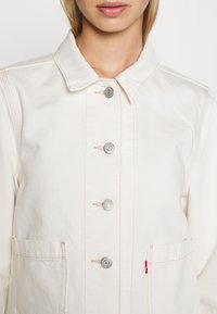 Ljus krämfärgad denimjacka med spetsig krage, fem silverfärgade tryckknappar, två framfickor och kontrasterande bruna sömmar.