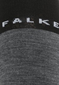 Calcetín gris con una textura acanalada, que presenta una parte superior negra. El nombre de la marca "FALKE" está impreso en blanco a lo largo del dobladillo.