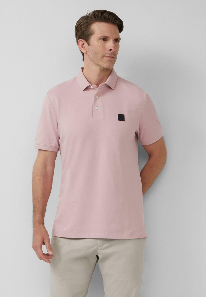 Polo rose clair en mélange de coton, avec un col classique, trois boutons, et un patch logo noir sur le côté gauche de la poitrine.