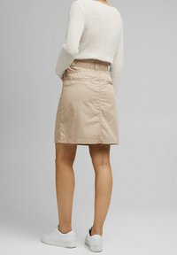 Jupe beige arrivant au genou avec une taille élastique, en tissu léger, dotée de deux poches arrière, associée à des baskets blanches.