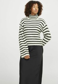 Pull à col roulé rayé en blanc cassé et noir, avec des manches lâches et une texture de tricot épaisse. Associé à une jupe noire.