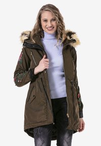 Olivgrüne Parka mit einer Kapuze aus Kunstfell, mit Reißverschlusstaschen und floraler Stickerei an den Ärmeln. Strukturiert mit matter Oberfläche.