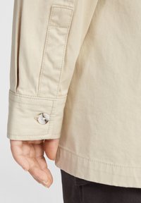 Tela de chaqueta beige con un acabado texturizado, que presenta un puño abotonado, costuras visibles y una abertura lateral cerca del dobladillo.