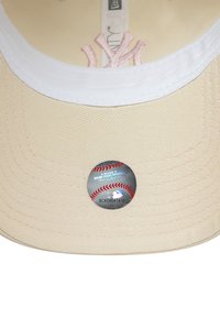 Bēša beisbola cepure ar balto izšūto logotipu un rozā adījumu. Tajā ir apaļš MLB uzlīme uz cepures malas. Gluda auduma tekstūra.