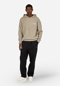 Světle hnědá chlupatá mikina s přední kapsou a bílým logem, doplněná volnými černými kalhotami a bílými teniskami, stojící na jednobarevném pozadí.