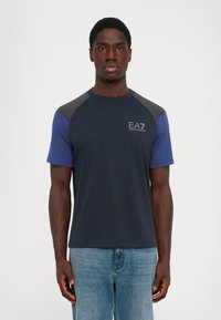 Vīrietis ar tumši zilu un pelēku īsām piedurknēm T-kreklu ar "EA7 Emporio Armani" logotipu uz krūtīm un gaiši zilām džinsām, stāv taisni.