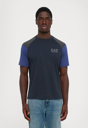 Man wearing a navy blue and gray short-sleeve t-shirt with "EA7 Emporio Armani" logo on the chest and light blue jeans, standing straight.