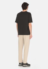 T-shirt nera in cotone con vestibilità rilassata, abbinata a pantaloni della tuta beige. L'insieme presenta sneakers grigie e nere, evidenziando un look casual.