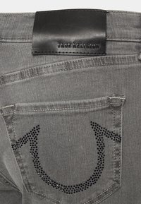 Vaqueros de mezclilla gris con una parche de cuero negro de la marca. Presentan un diseño destacado de piedras brillantes en forma de herradura en los bolsillos traseros.