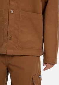 Braune Baumwolljacke mit Knopfverschlüssen, zwei Vorderaschen und glatter Textur, kombiniert mit passenden braunen Hosen und einer Cargo-Tasche.