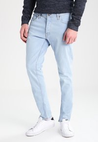 Vaqueros de mezclilla azul claro con corte ajustado, cinco bolsillos y cierre de botones. Combinados con zapatillas blancas para un look casual.