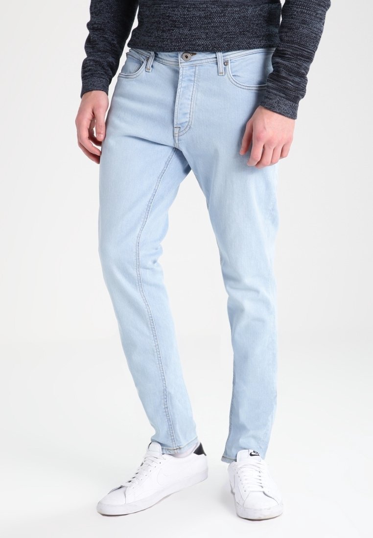 Vaqueros de mezclilla azul claro con corte ajustado, cinco bolsillos y cierre de botones. Combinados con zapatillas blancas para un look casual.