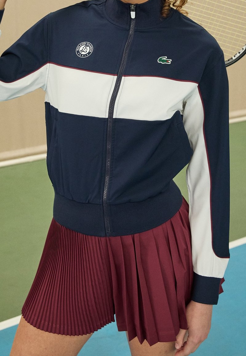 Mörkblå zip-up jacka med vita och vinröda ränder samt Lacoste-logotyp, i kombination med en vinröd veckad kjol. Slät textur, sportig design.