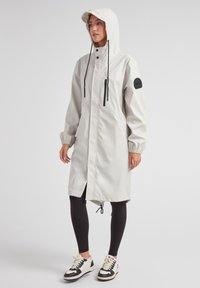 Cappotto impermeabile grigio chiaro con cappuccio, tasca sul petto con zip, polsini elasticizzati e orlo con cordino, abbinato a leggings neri e sneakers.