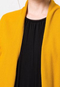 Cardigan amarillo texturizado con frente abierto, combinado con una blusa negra de pliegues. El cardigan presenta un patrón de panal y un ajuste holgado.