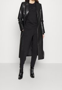 Svart läder lång jacka över svart tröja och byxor, med en subtil grafisk detalj, parat med eleganta svarta ankelskoot.