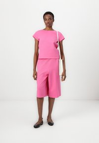 Ροζ βαμβακερό σετ δύο κομματιών με κοντομάνικο τοπ και σορτς μέχρι το γόνατο. Το μοντέλο κρατάει μια λευκή τσάντα και φοράει μαύρες φλατ παντόφλες.