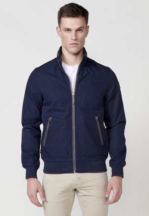 Chaqueta de cremallera azul marino con collar alzado, dos bolsillos frontales con cremallera y puños de canalé. Presenta un logotipo en el pecho y un tejido texturizado.