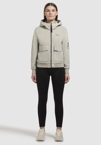 Light grey zip-up jacket with a hood, two front pockets, ribbed cuffs, and an embroidered logo on the chest; paired with black leggings and sneakers.