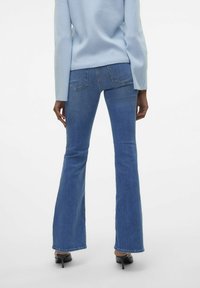 Modre kavbojke s širokimi nogavicami in srednjim pasom, z zadnjimi žepi in gladko, rahlo strukturirano površino. Ujemajo se z črnimi visokimi petami.