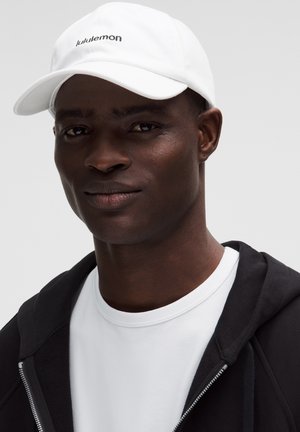 Man met witte lululemon-pet, wit T-shirt met ronde hals en een zwart openritsbare hoodie tegen een lichtgrijze achtergrond.