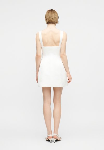 Femme debout, de dos, portant une mini robe blanche sans manches et des sandales blanches à talons bas sur un fond clair uni.