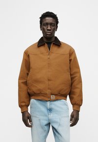 Άνδρας που φοράει καφέ μπουφάν με φερμουάρ και σκούρο γιακά και ανοιχτό μπλε τζιν, στέκεται μπροστά σε απλό λευκό φόντο.