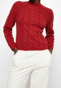 Personne portant un pull en tricot rouge à manches longues et un pantalon blanc taille haute, les mains dans les poches.