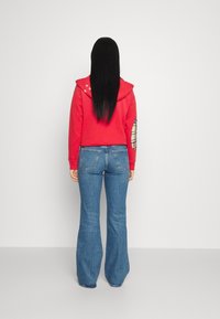 Červená mikina s kapucí na stahování a dekorativními hvězdami; doplněná modrými džínami s rozšířenými nohavicemi a hadříkem v káro na jedné kapse.